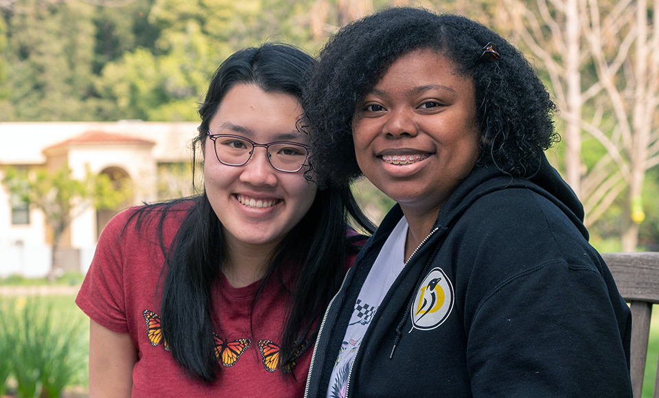 two female students sitting together on a bench on the ֱ campus smile at the camera