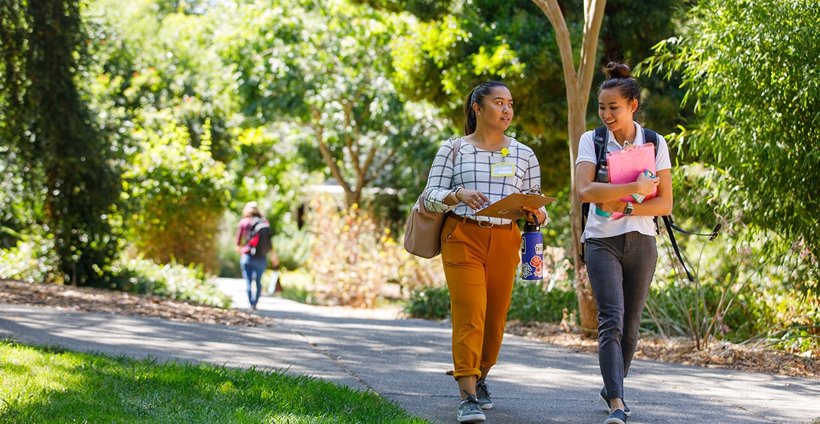 two students walk down a path with their books and backpacks on the ֱ campus