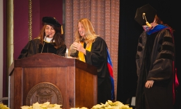 Photo of Lynn Sondag (center) in regalia receiving Fink Faculty Award at podium during 2022 Fall Commencement ceremony flanked by Mojgan Bermand (left) and ֱ President Nicola Pitchford