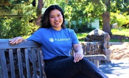 Christina Pathoumthong, wearing blue Fulbright t-shirt, sits on gray wooden bench on stone rock bridge on ֱ campus