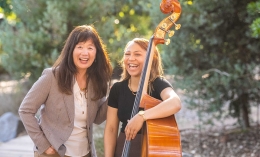 ֱ University of California Chair of Music Department, June Oh and a female student holding a bass are outdoors on the ֱ campus. Both women are laughing.