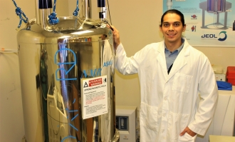 Photo of ֱ adjunct professor Joseph Morris '19 '21 MS standing on right wearing white lab coat with his right hand on placed on top of silver NMR spectrometer