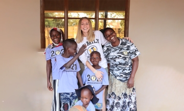 Photo of student-athlete Jordan Charlton standing wearing white ֱ Athletics long-sleeved T-shirt posing and smiling with arms around young Ugandan students smiling and wearing gray ֱ athletics T-shirts
